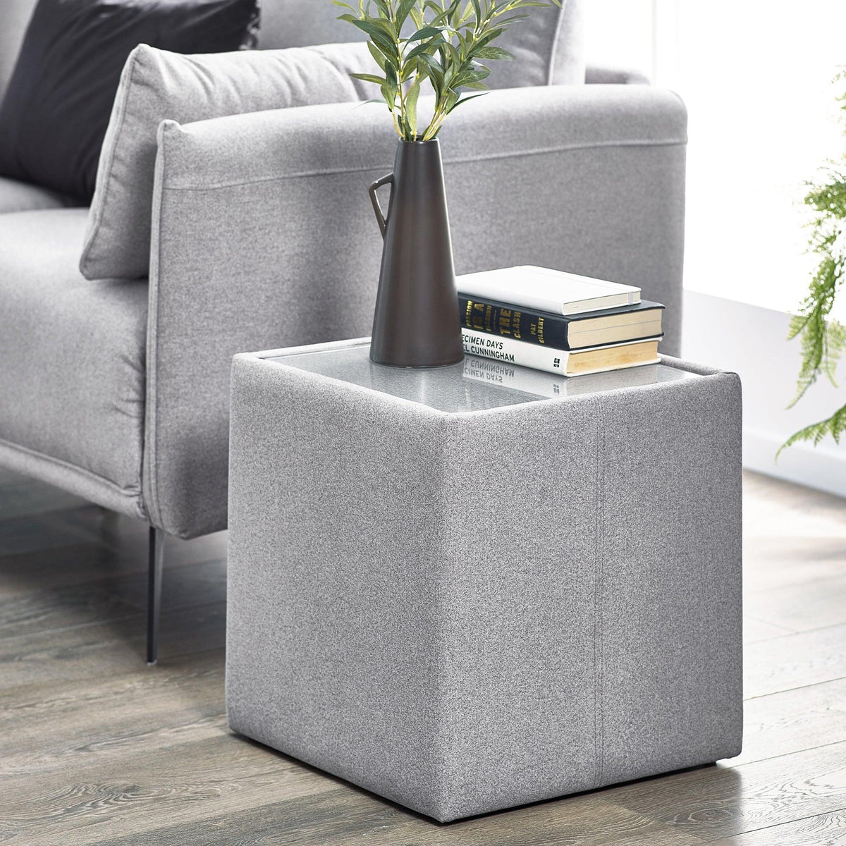 Grey fabric lamp table with glass top beside sofa, books and vase on surface