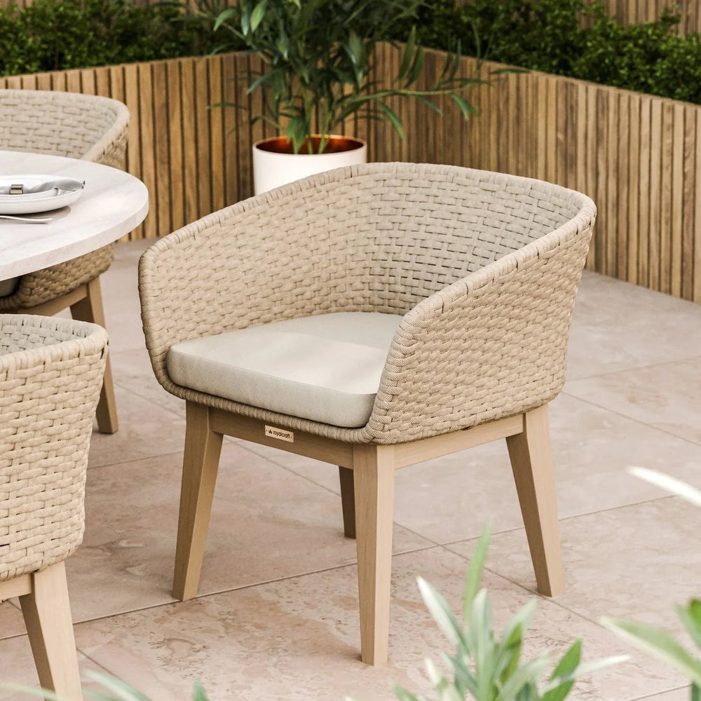 Beige woven outdoor dining chair with cushion on tiled patio, wooden planter in background