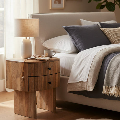 Wooden nightstand with lamp and mug next to a bed with pillows and blanket