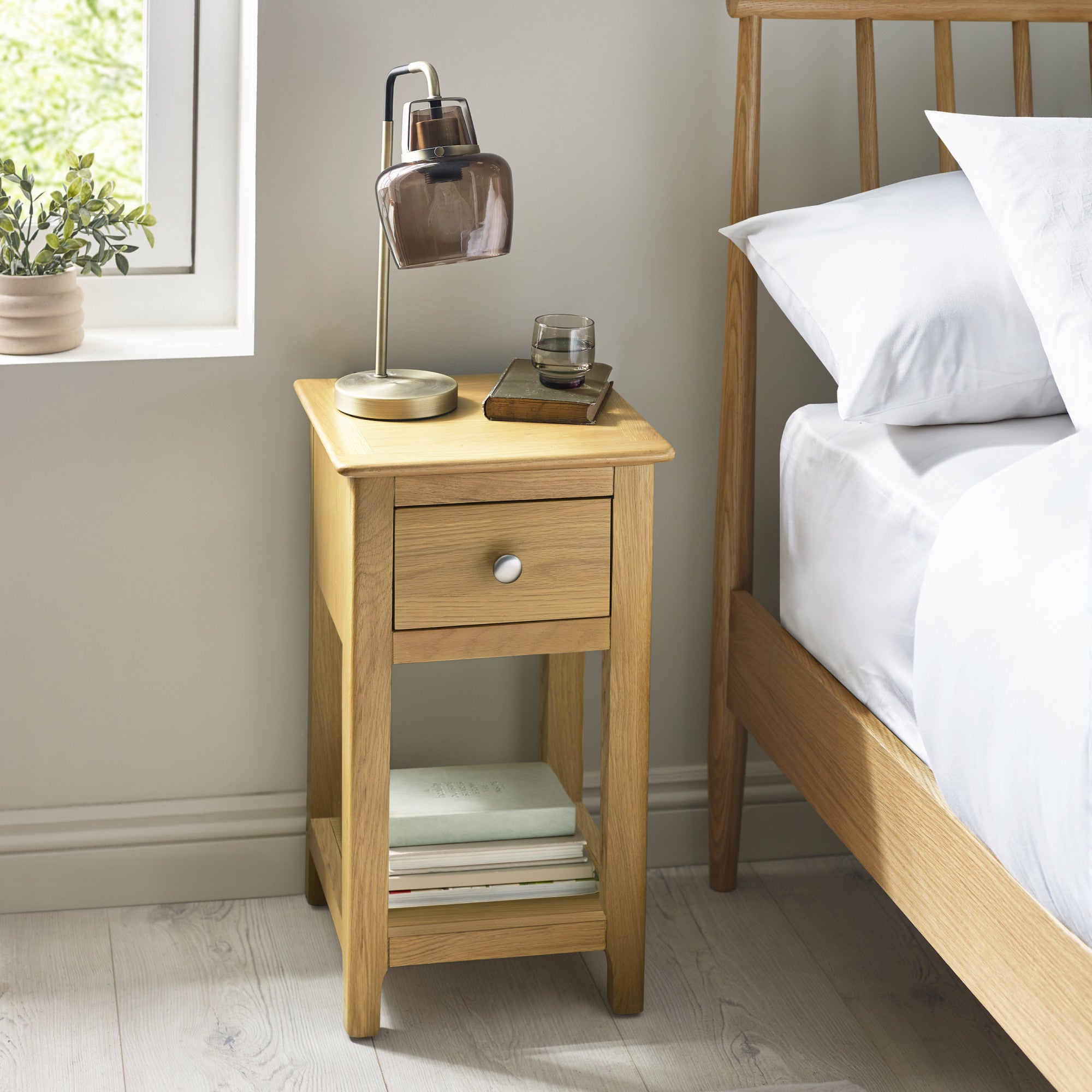 Light oak bedside table with single drawer and open shelf, shown in bedroom setting with lamp and books