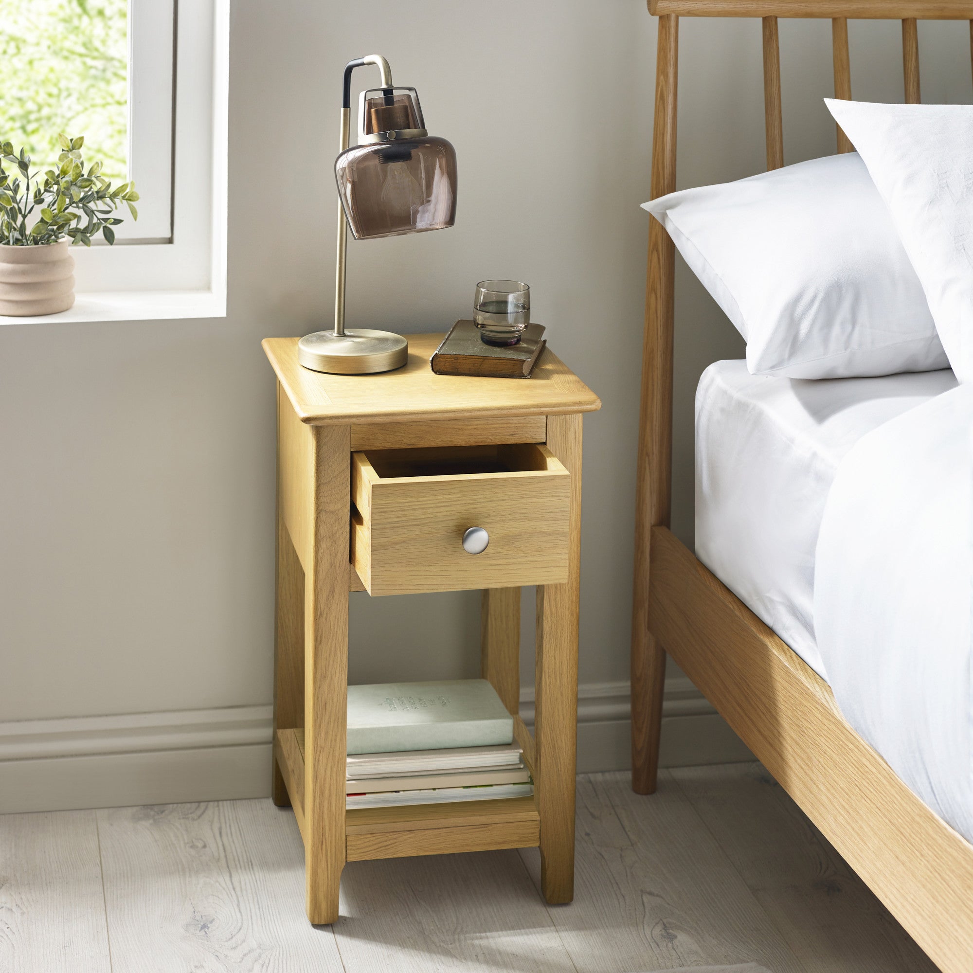 Light oak bedside table with one open drawer, glass lamp, book, and glass on top beside bed