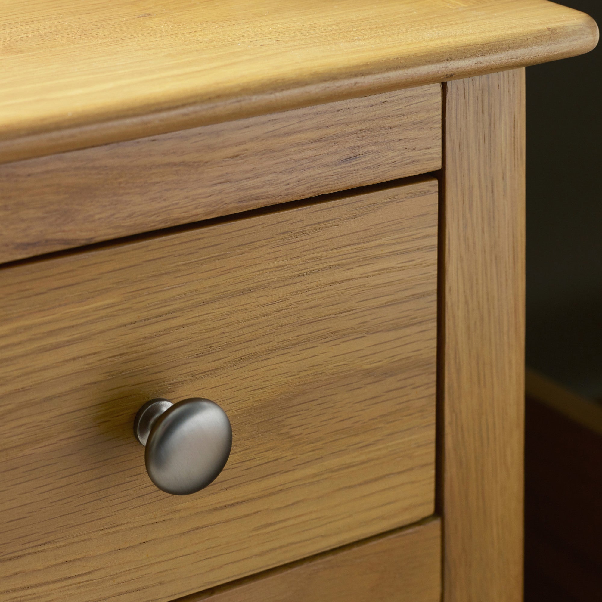 Close up of silver metal knob on light oak bedside table drawer, showing wood grain texture