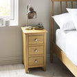 Light oak 3 drawer bedside table in bedroom setting with white bed and table lamp on top