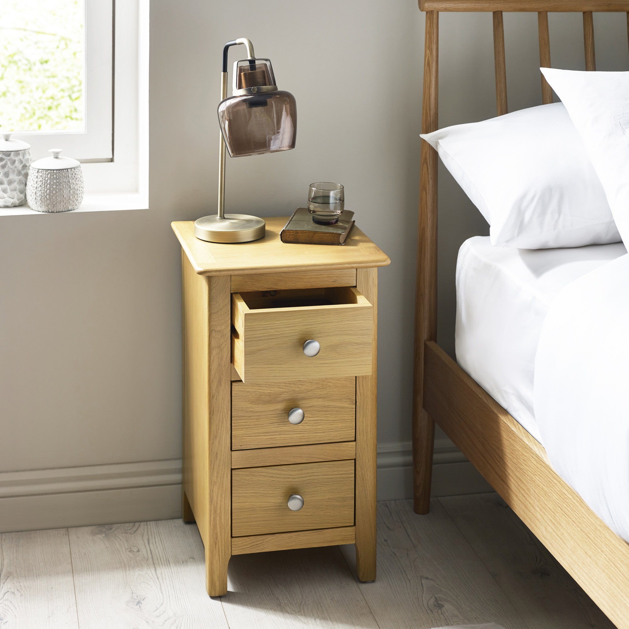 Same bedside table shown with top drawer partially open, placed next to a wooden bed frame