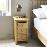 Same bedside table shown with top drawer partially open, placed next to a wooden bed frame