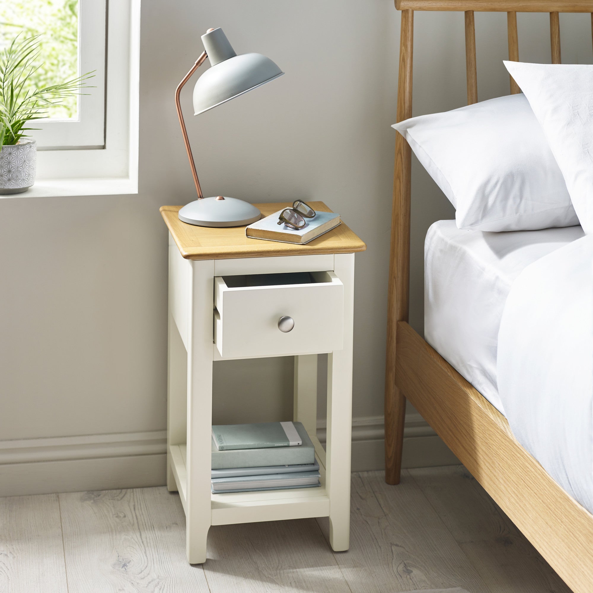 Cream painted bedside table with oak top and one drawer partially open beside bed in room