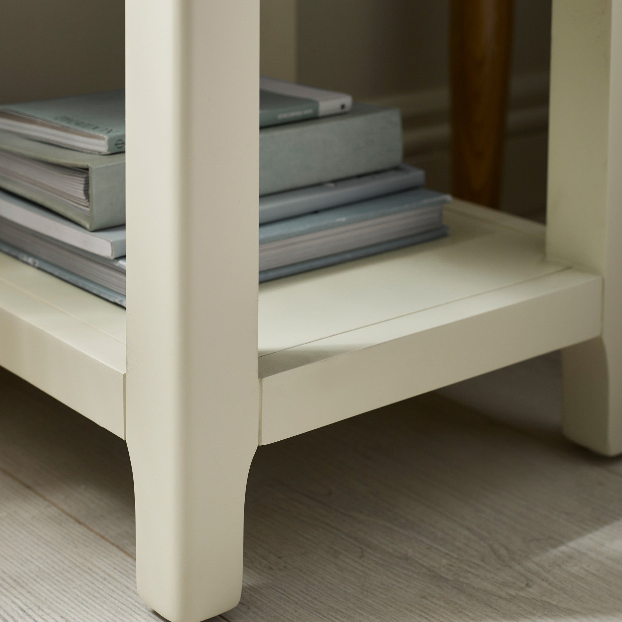 Close-up of cream painted bedside table leg with shelf holding stacked books on wood floor