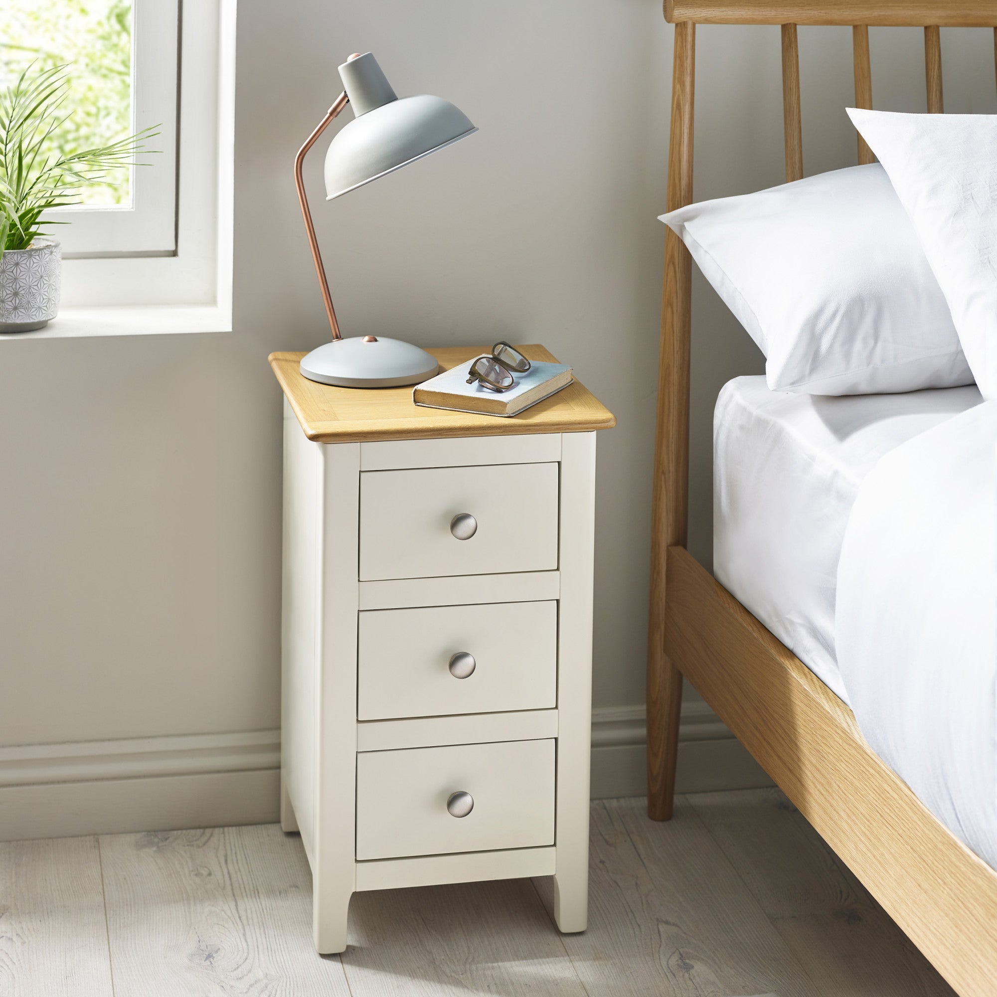 Cream colored 3 drawer bedside with oak tabletop next to wooden bed and window, decorated with lamp, book, and glasses