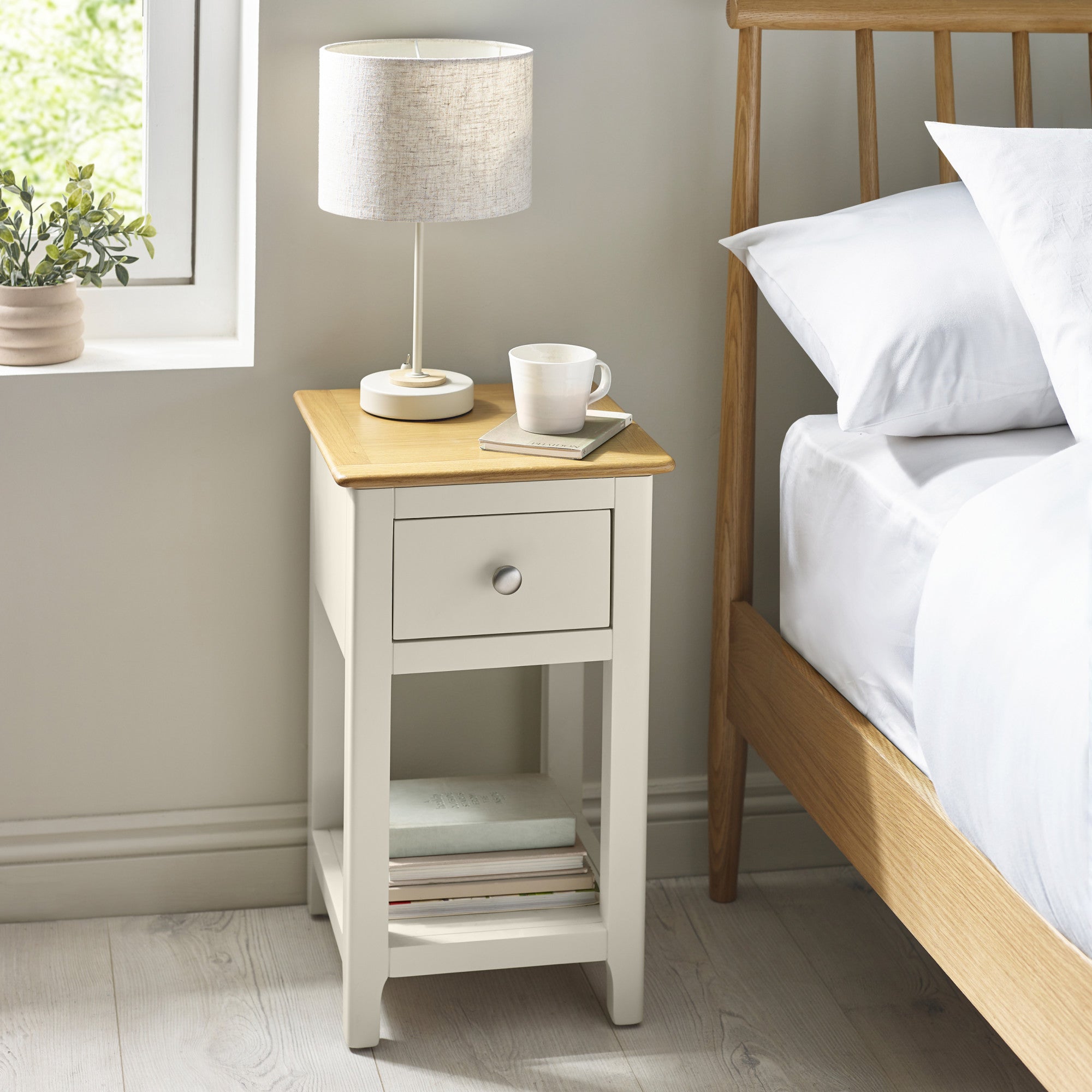 Bedside table next to bed with lamp, cup, and books, in a light and neutral bedroom