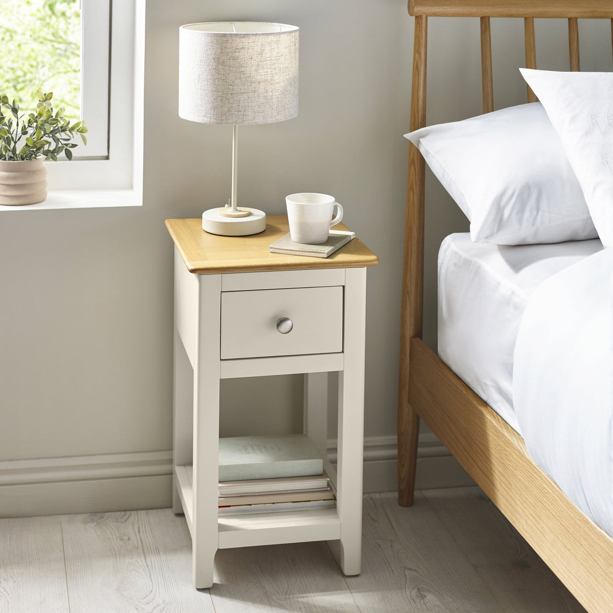 Bedside table next to bed with lamp, cup, and books, in a light and neutral bedroom