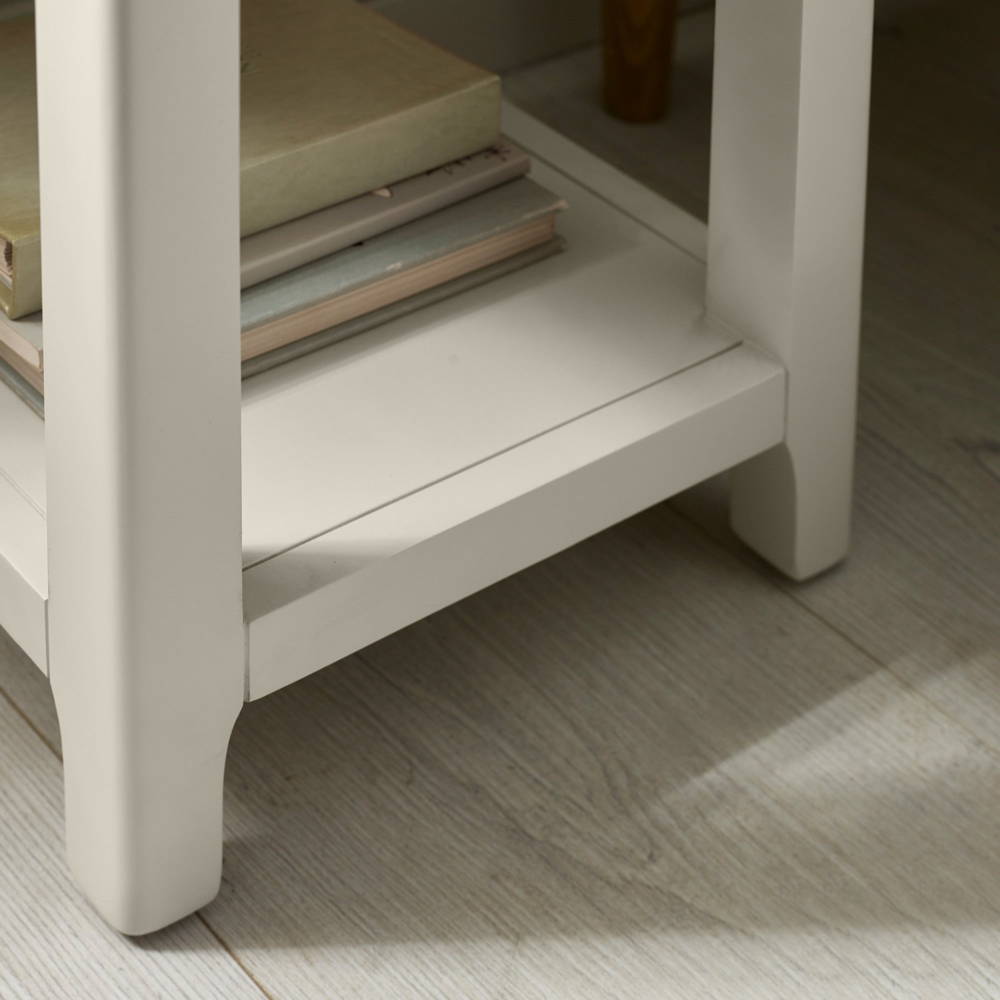 Close-up of bottom shelf and legs of light grey painted bedside table on wooden floor with books underneath.