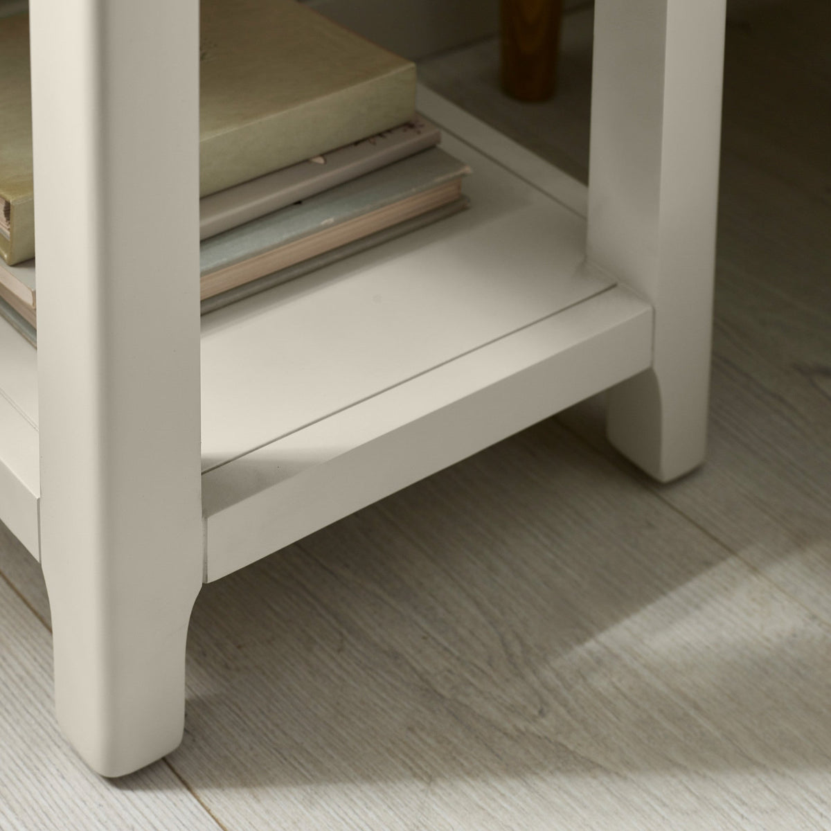 Close-up of bottom shelf and legs of light grey painted bedside table on wooden floor with books underneath.
