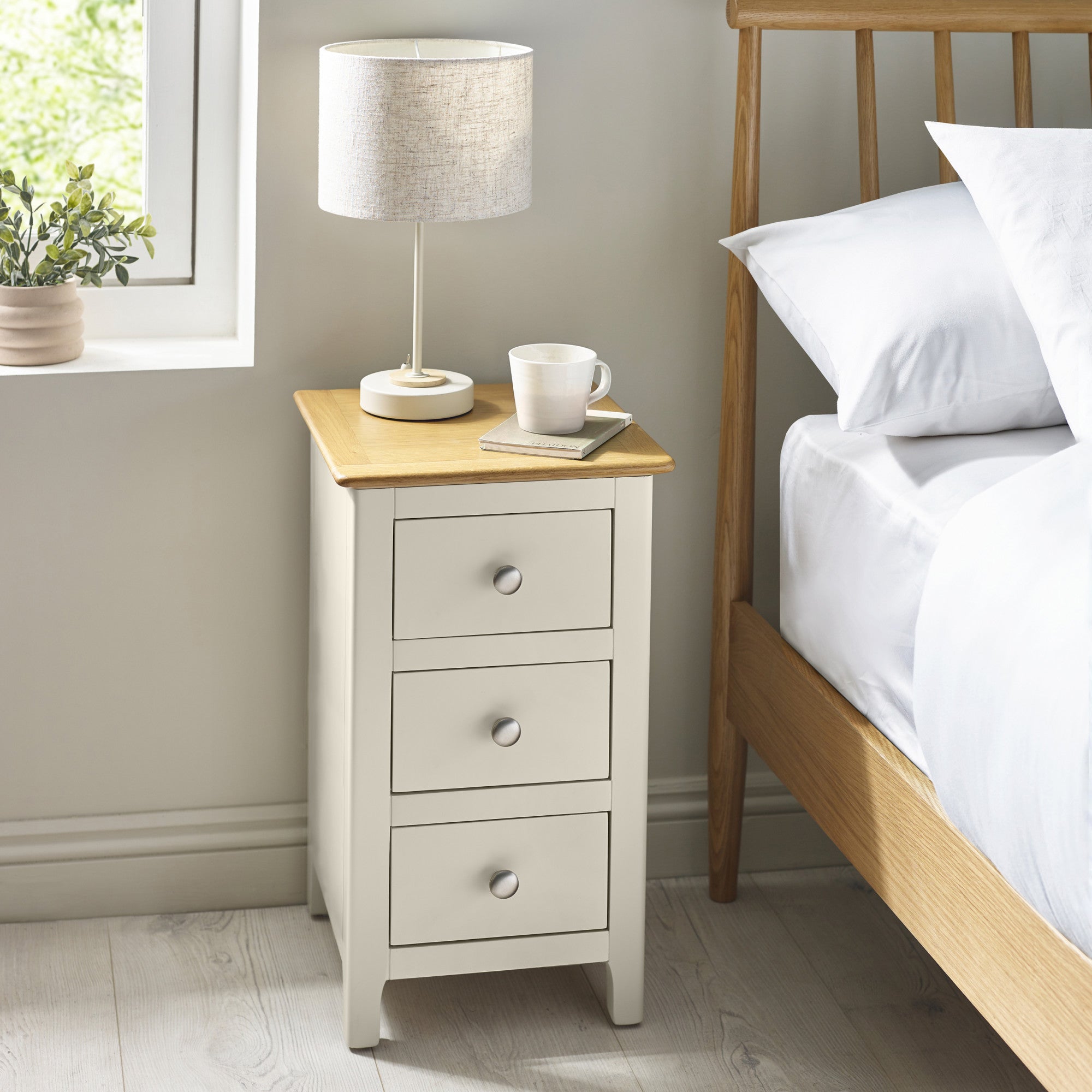 Beige 3 drawer bedside with lamp, book, cup on top, next to bed with white bedding in room