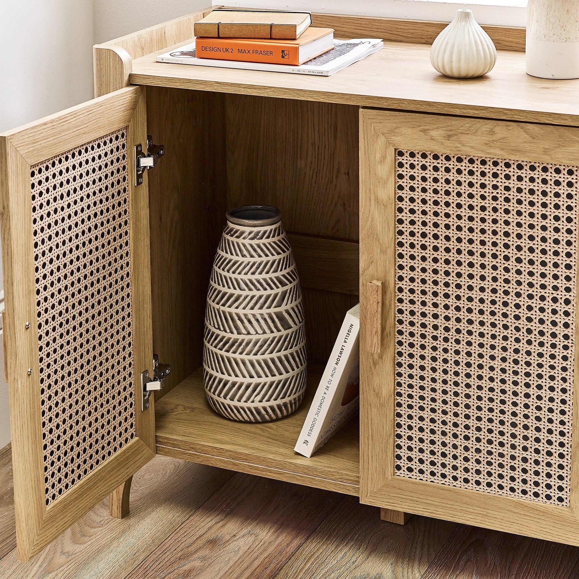 Rattan sideboard with open door showing storage shelf, ceramic vase, and wooden surface