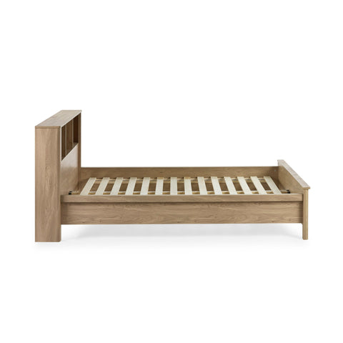 Side view of oak wood bed frame with slatted base and headboard shelving against white background