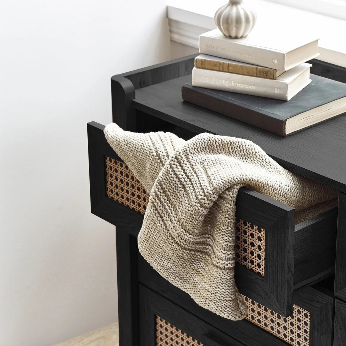 Black rattan effect wide chest with open drawer, knit blanket, stacked books and decor