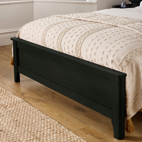 Black wooden bed footboard with a textured beige blanket in a room with wooden flooring and woven rug