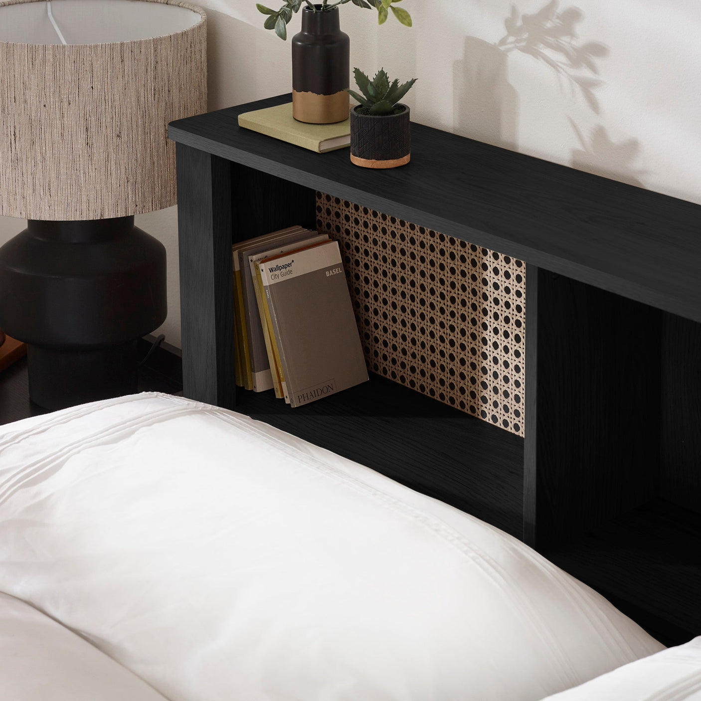Black wooden headboard with cane panel inset, decorated with books and plants, beside a lamp on a nightstand
