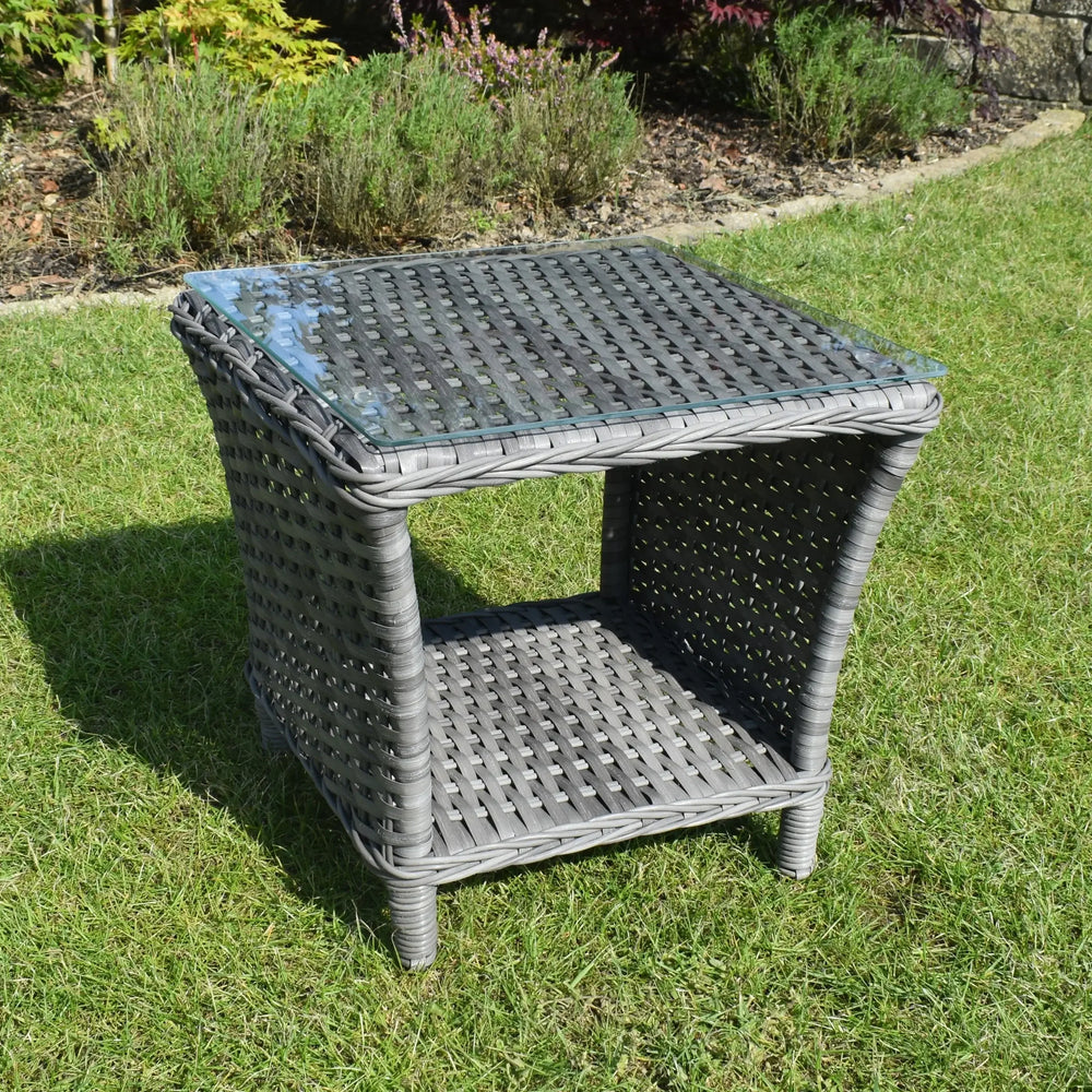 Grey rattan garden side table with glass top and storage shelf on grass