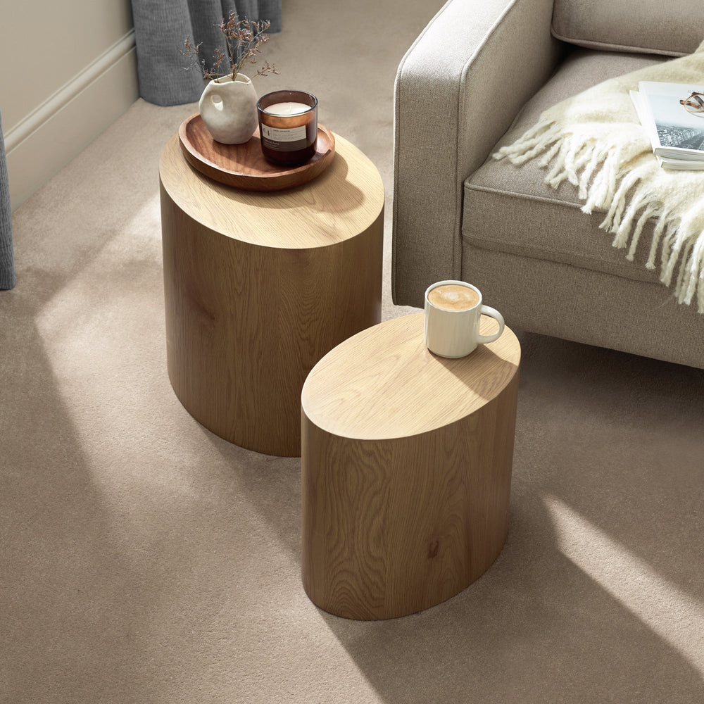 Two light oak oval nest tables on beige carpet beside a beige sofa, one with a coffee and the other with a candle and vase.