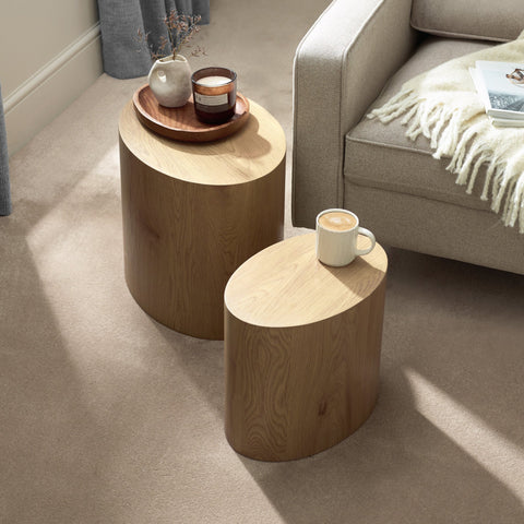 Two light oak oval nest tables on beige carpet beside a beige sofa, one with a coffee and the other with a candle and vase.