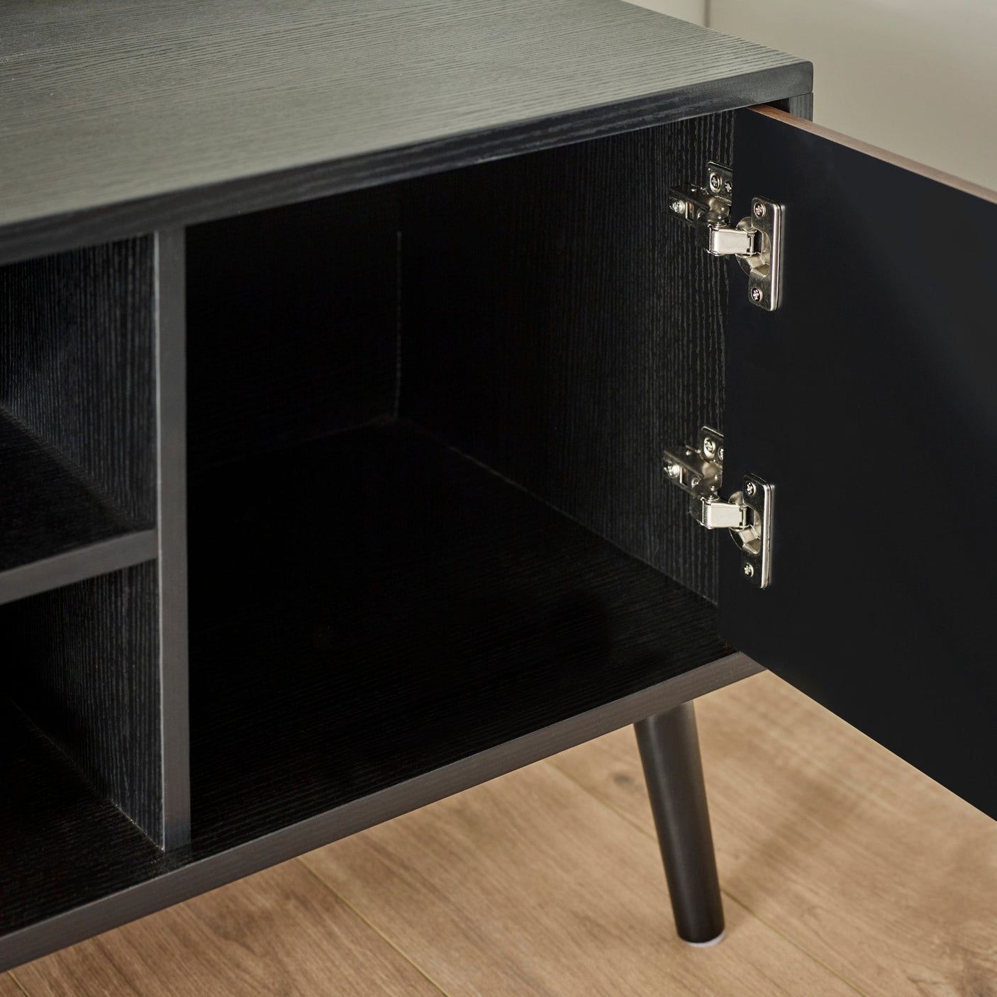 Open black cabinet door showing black wood interior with metal hinges inside TV unit on light wood flooring