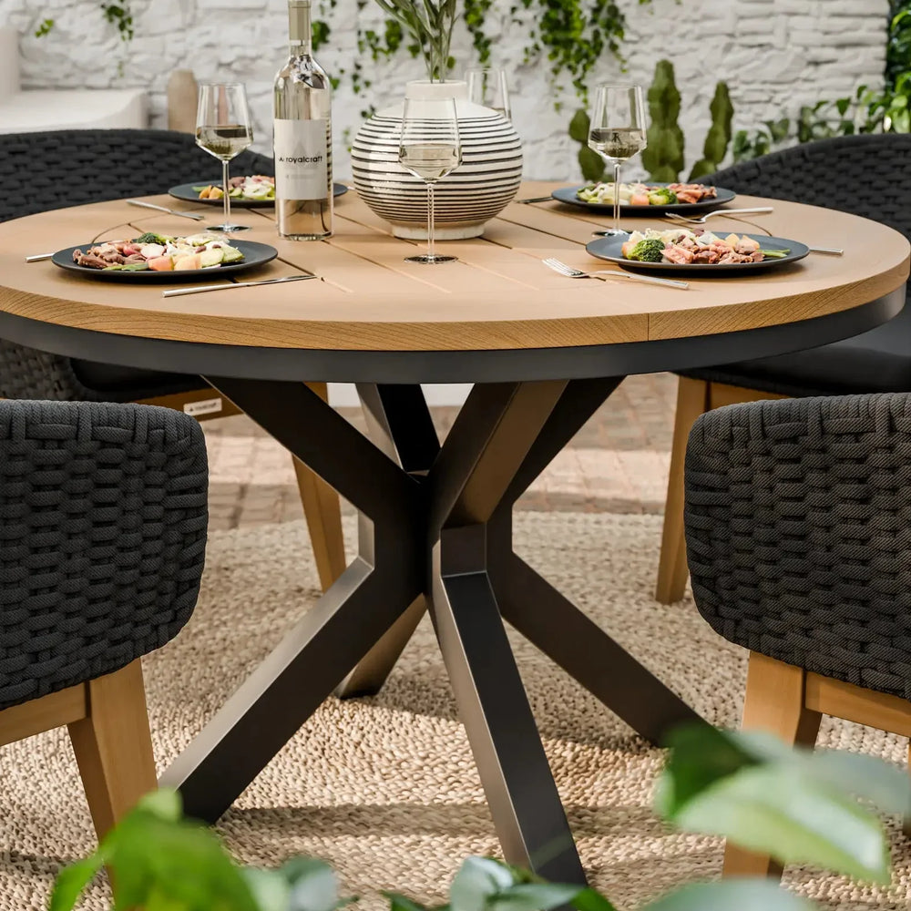 Round wooden dining table with black chairs set for four, outdoor setting with greenery