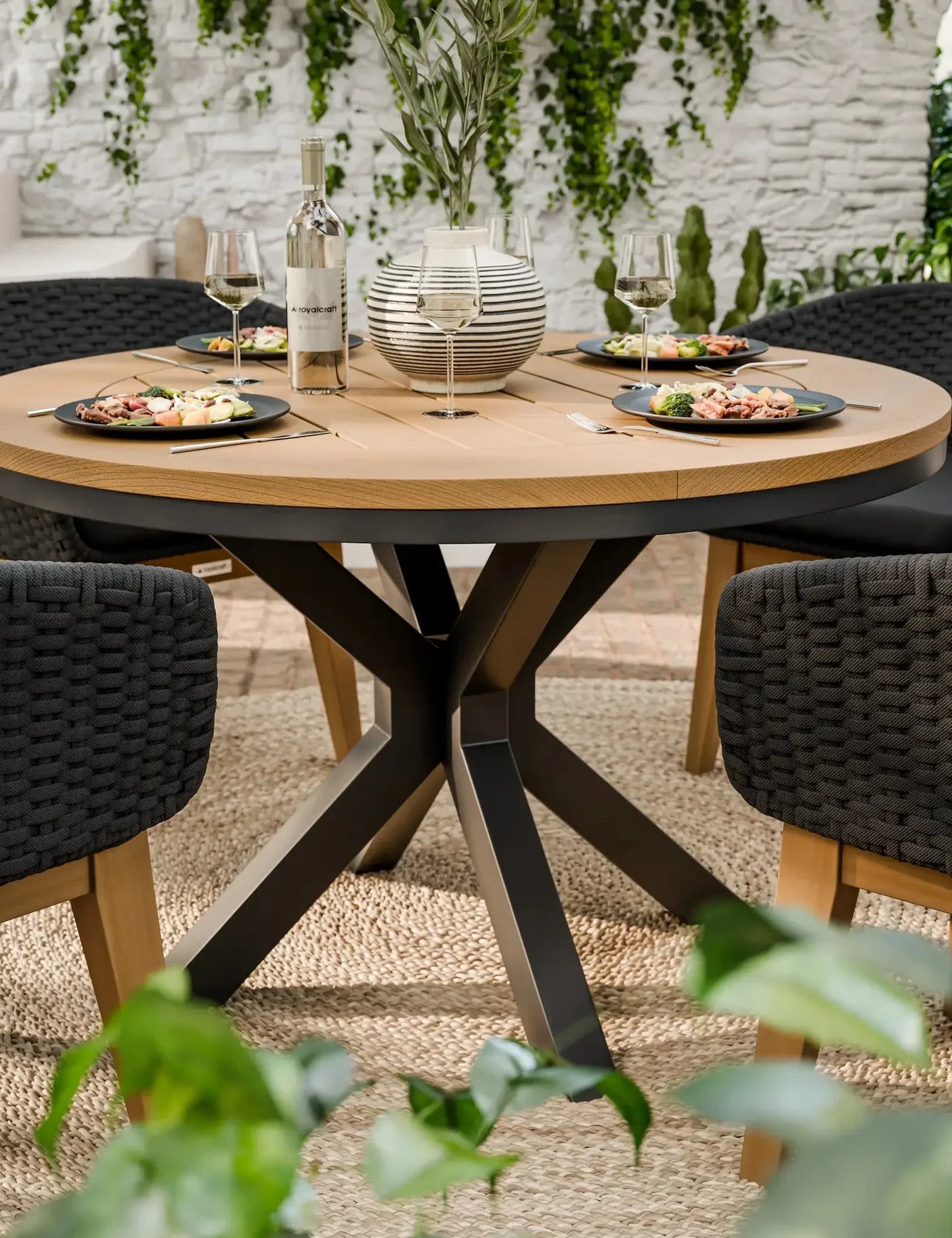 Round wooden dining table with black chairs set for four, outdoor setting with greenery