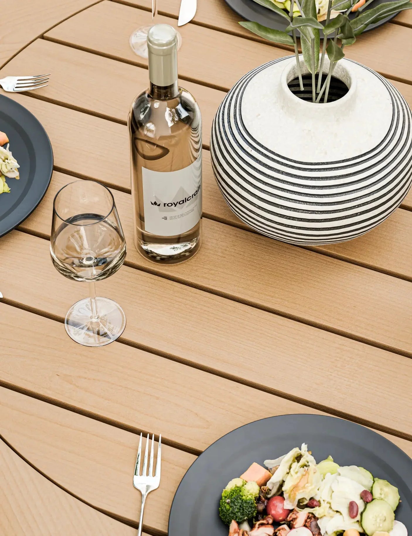 Wood effect outdoor dining table with plates, cutlery, glass, wine bottle and striped vase