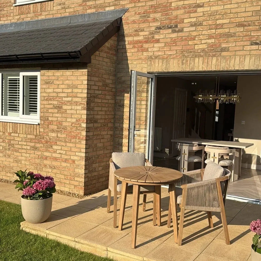Wooden outdoor dining set with three chairs on patio beside brick house and potted flowers