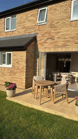 Wooden outdoor dining set with three chairs on patio beside brick house and potted flowers