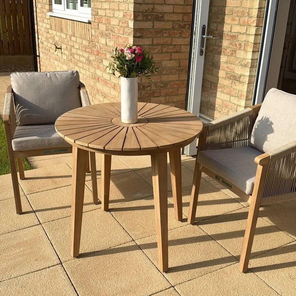 Wooden outdoor dining set with round table, two cushioned chairs, vase of flowers on patio