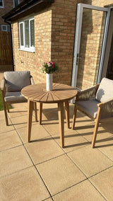 Wooden outdoor dining set with round table, two cushioned chairs, vase of flowers on patio