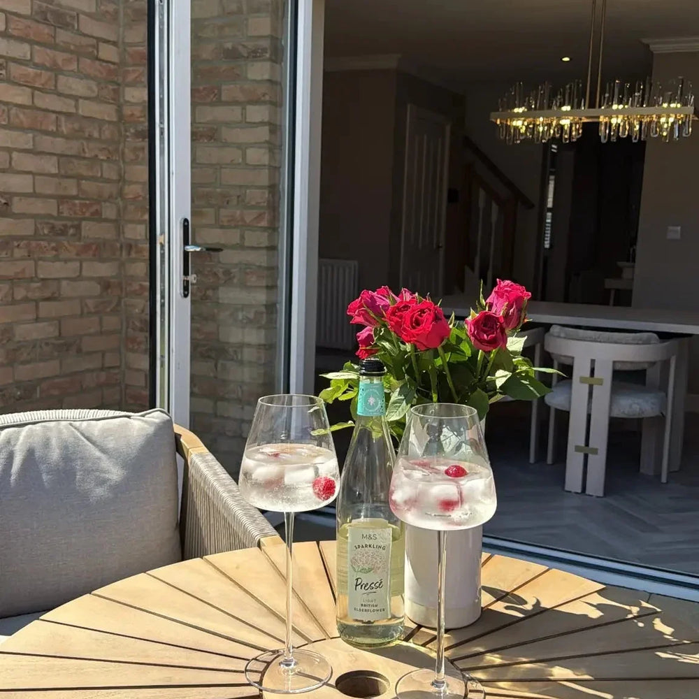 Round wooden garden table with two drinks, bottle, and vase of red roses on patio