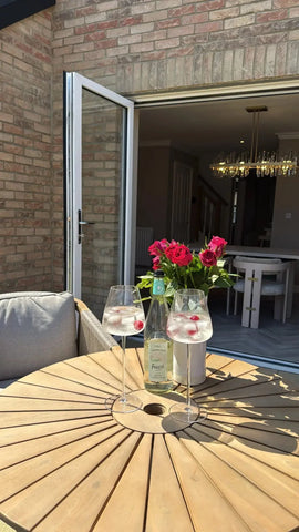 Round wooden garden table with two drinks, bottle, and vase of red roses on patio