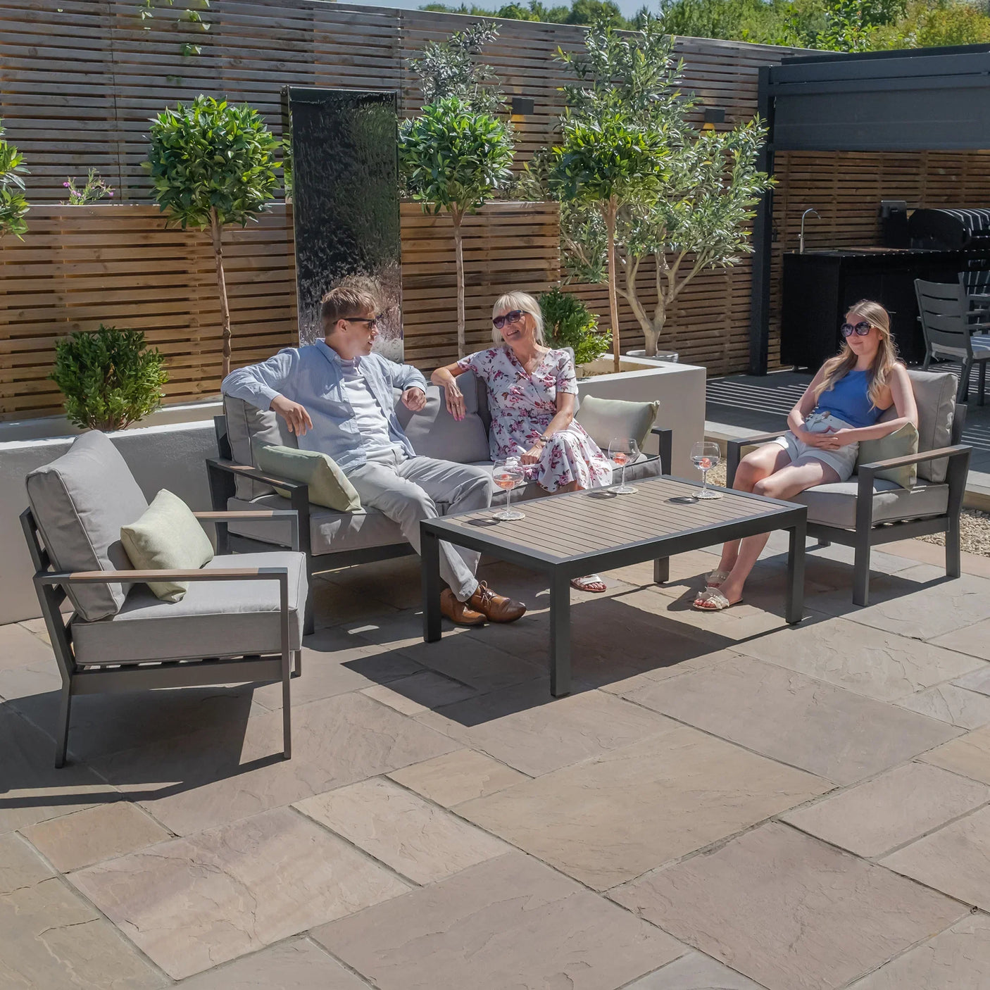 Grey outdoor sofa set with armchairs and coffee table on patio, people relaxing in sunlight