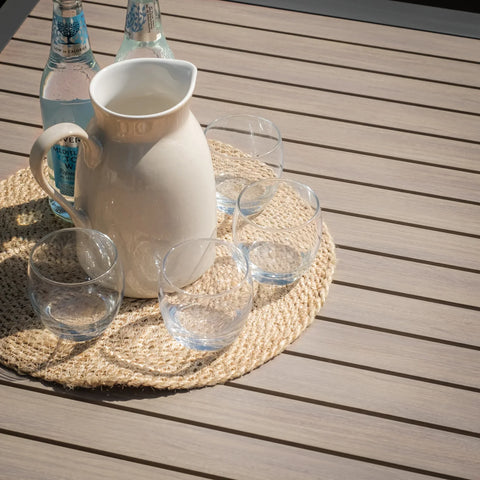 Light wood outdoor dining table with jug, glass tumblers, and water bottles on woven mat