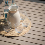Light wood outdoor dining table with jug, glass tumblers, and water bottles on woven mat