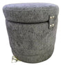 A round, upholstered storage ottoman or stool with a textured gray fabric cover, featuring a small side tab with gold-tone metal studs and a small gold tag near the base, shown on a plain white background.