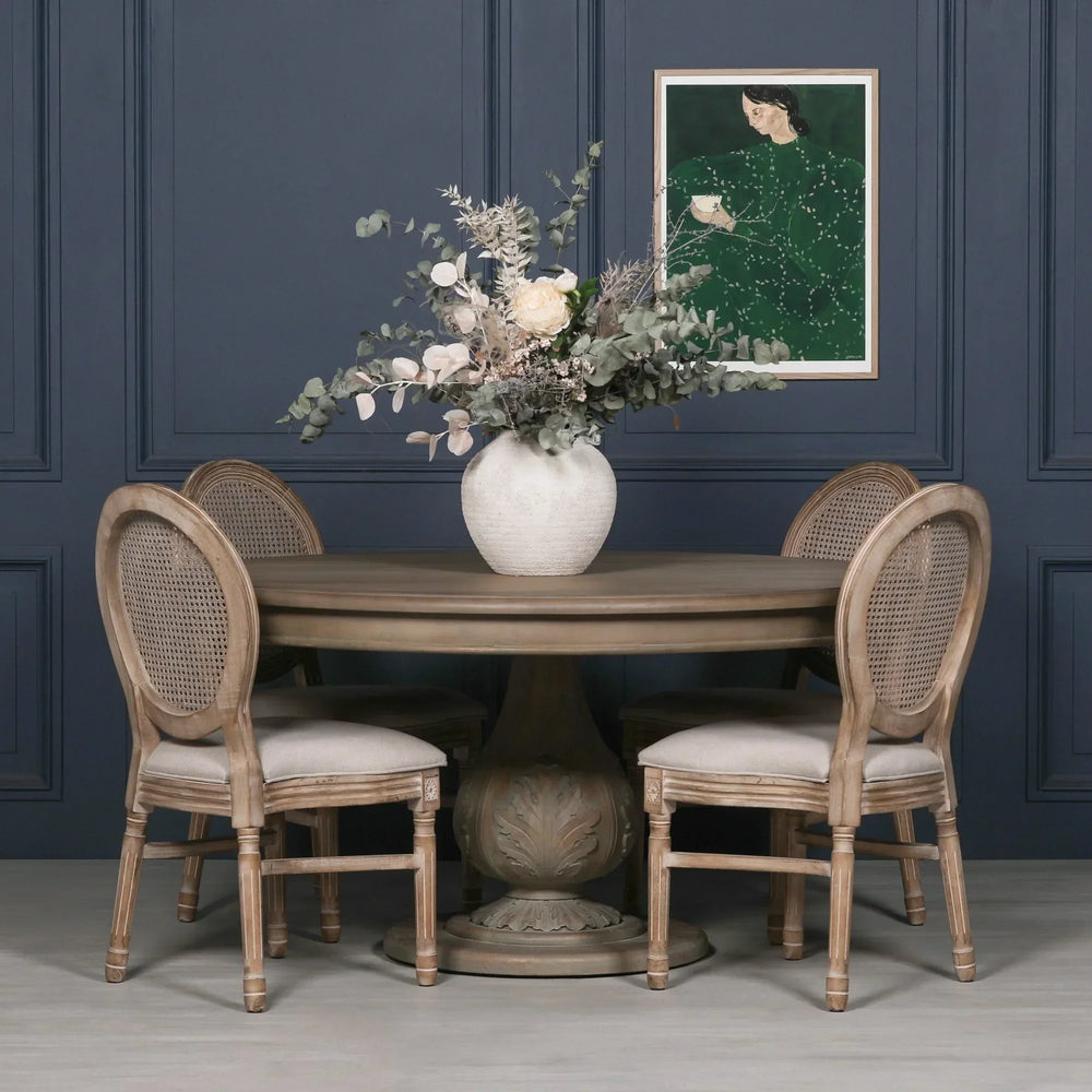 Round oak dining table with carved pedestal base and four rattan back chairs, floral centerpiece
