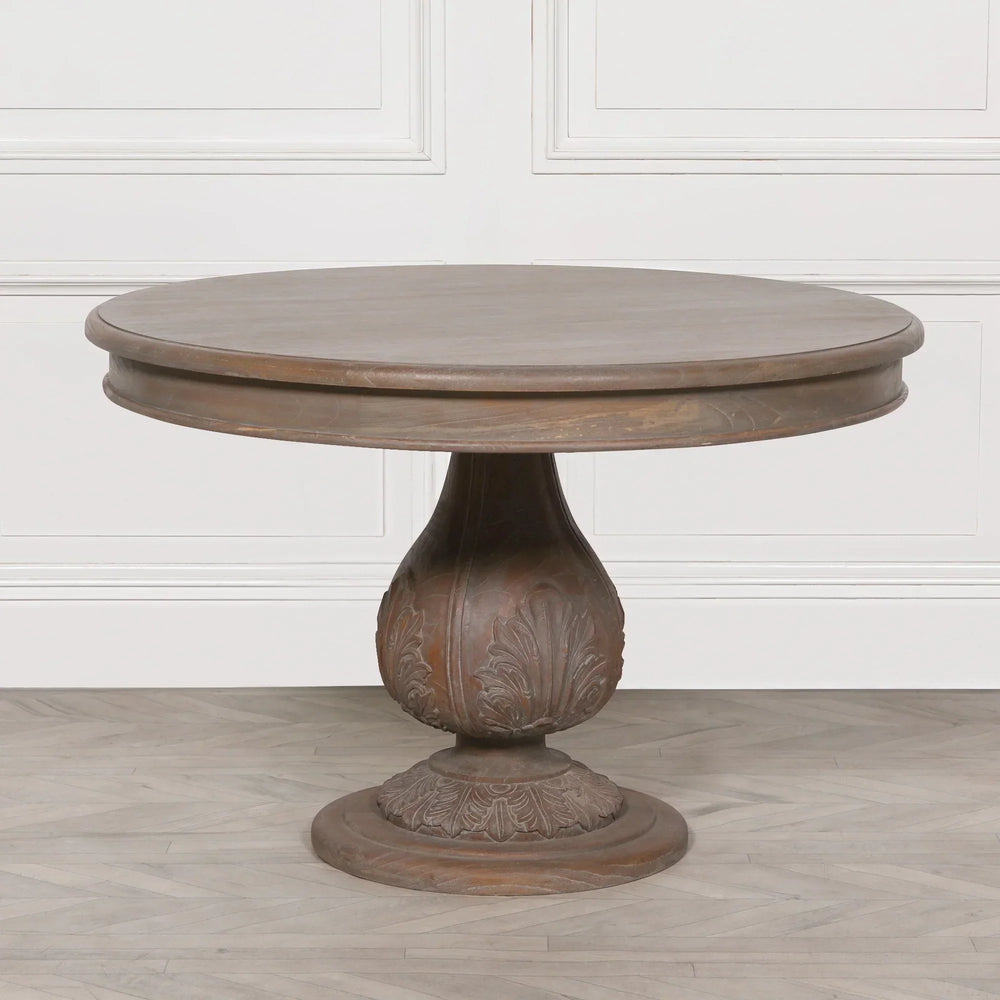Round wooden dining table with carved pedestal base on light floor