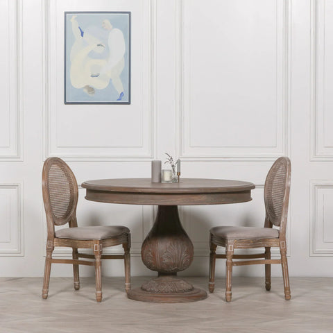 Round wooden dining table with pedestal base and two cane back chairs in neutral room