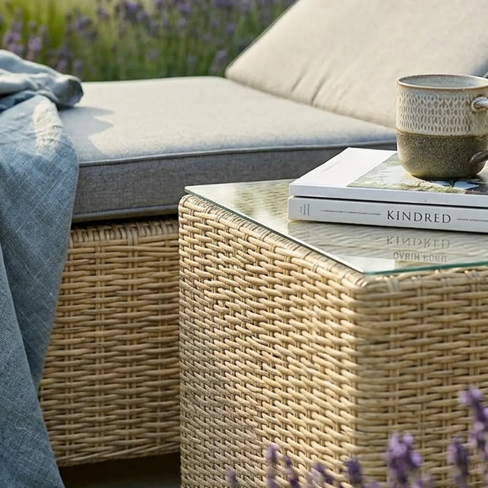 Rattan outdoor coffee table with glass top, books, and mug beside grey cushioned garden sofa