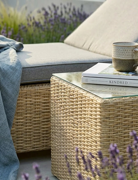 Rattan outdoor coffee table with glass top, books, and mug beside grey cushioned garden sofa