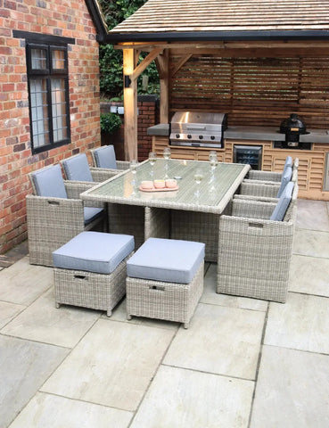 Rattan garden dining set with glass top table and grey cushions on a patio near outdoor kitchen
