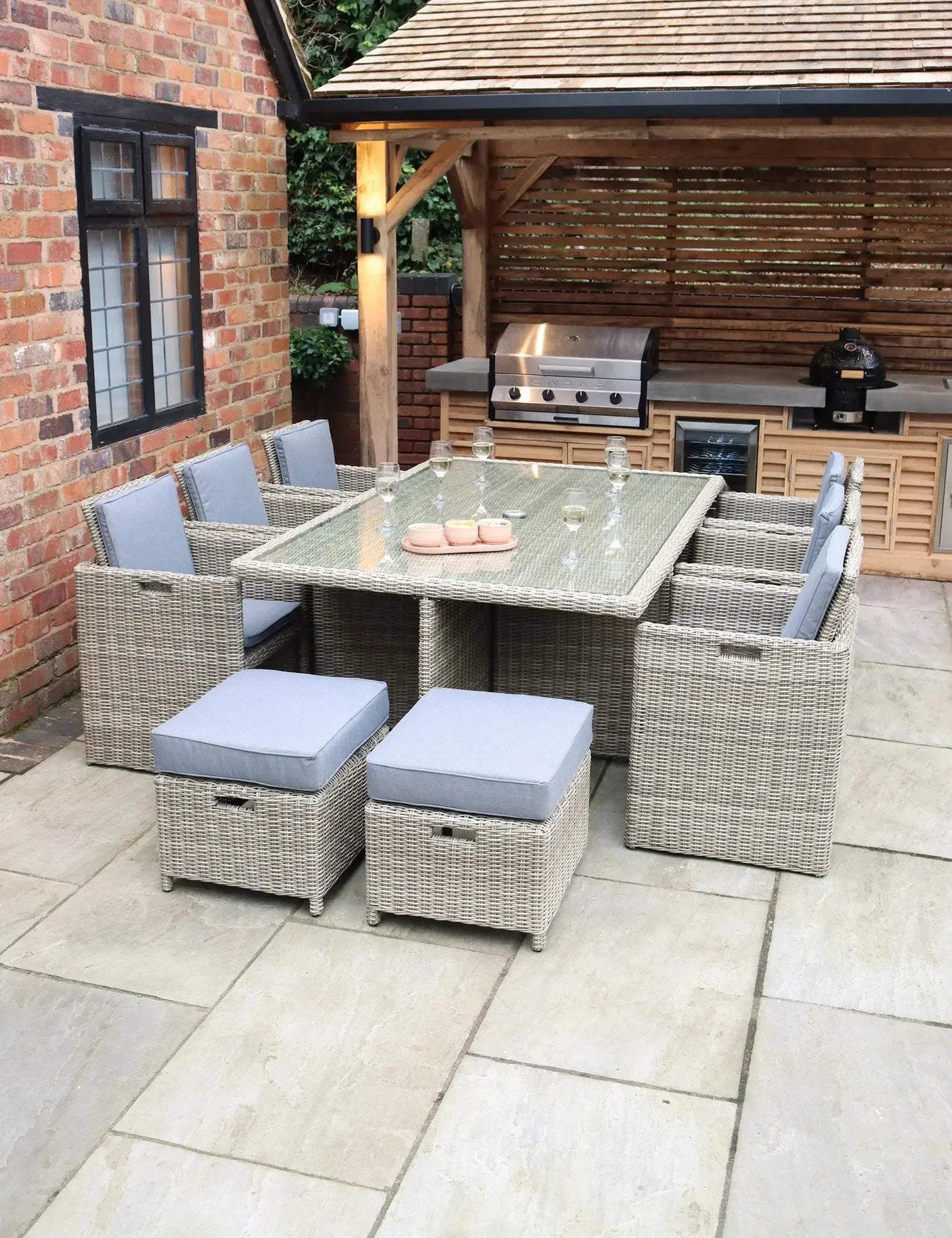 Rattan garden dining set with glass top table and grey cushions on a patio near outdoor kitchen