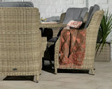 Rattan dining set with grey cushions and throw, set against a white brick wall with plant decor
