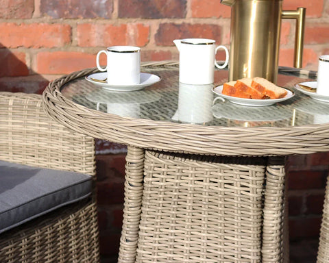 Rattan garden table set with glass top, two cups, plates, and a brass jug outdoors