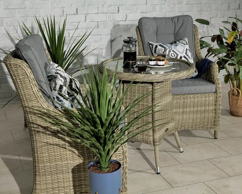 Rattan bistro set with glass table, grey cushions, and patterned pillows in indoor setting