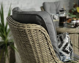 Close up of rattan garden chair with grey cushion and printed pillow, glass table in background